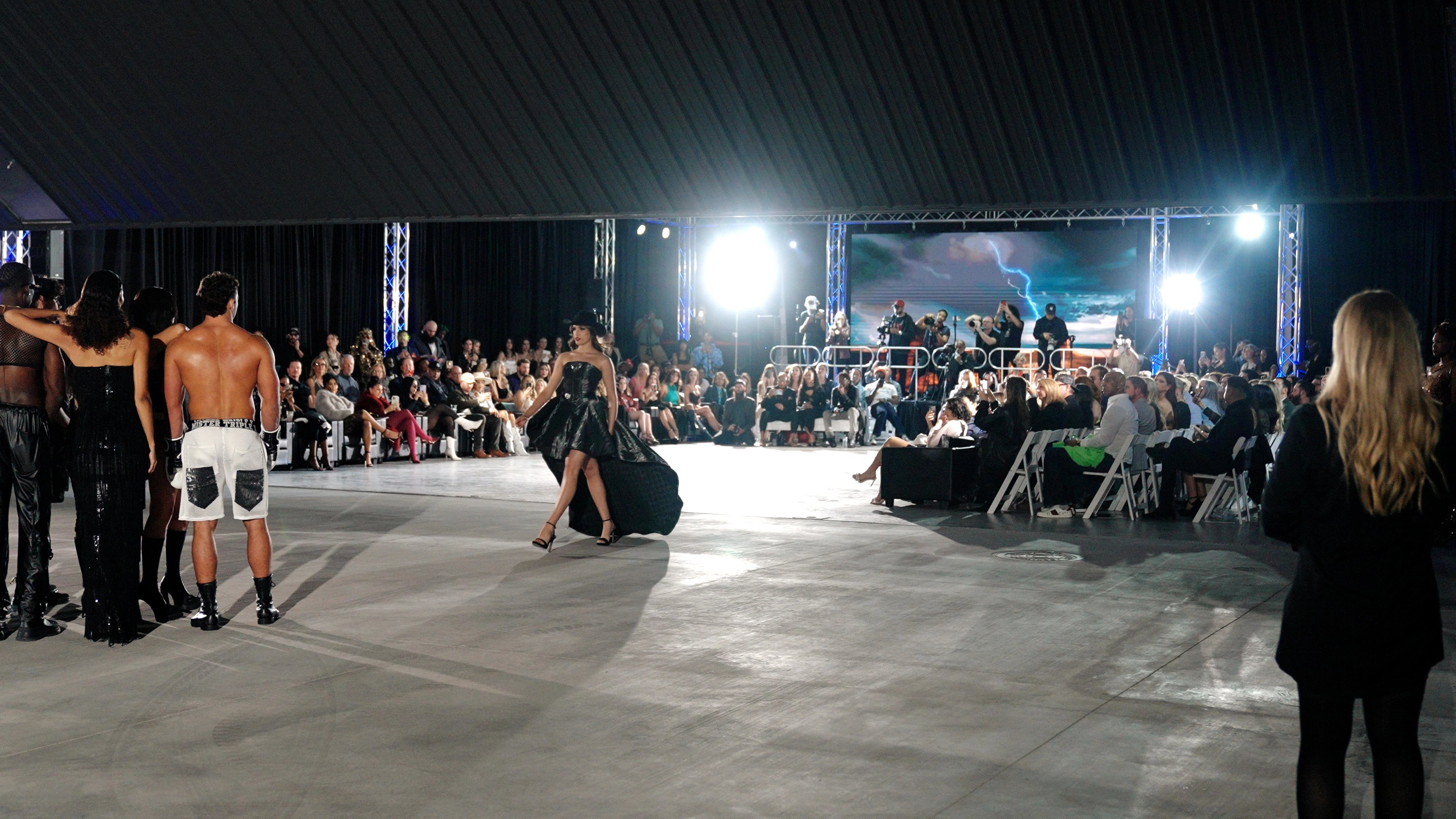 Model walking an open-air LaCreme runway in a black avant-garde dress in front of a seated audience and photographers.